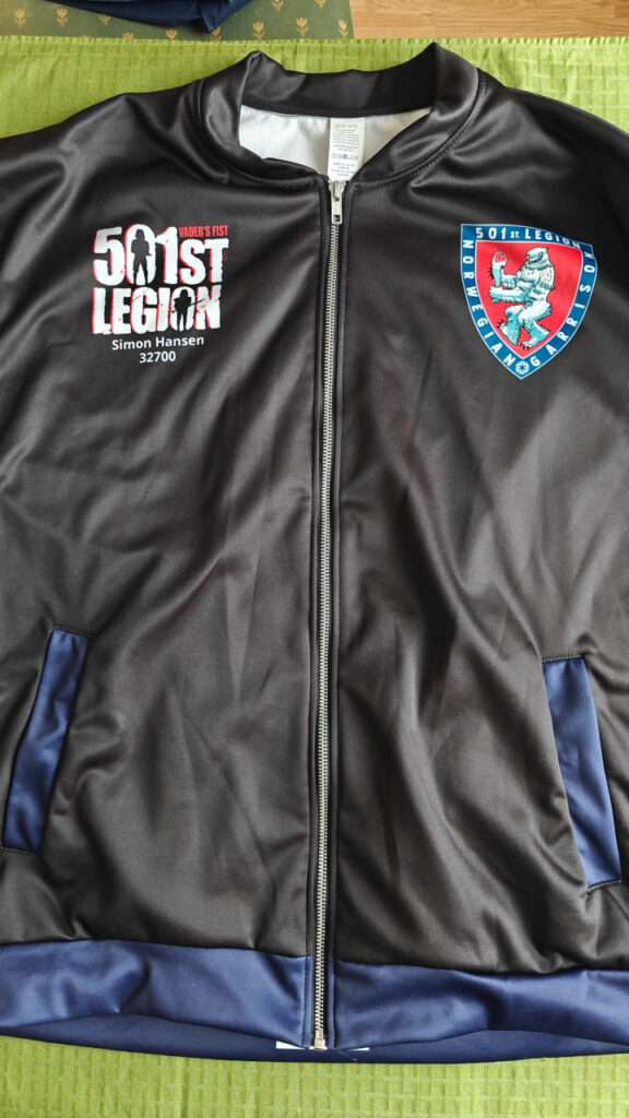 A black nylon jacket with blue pockets. The 501st letter logo and my name and ID number is on the right side of the chest, and the garrison logo is on the left side of the chest. 