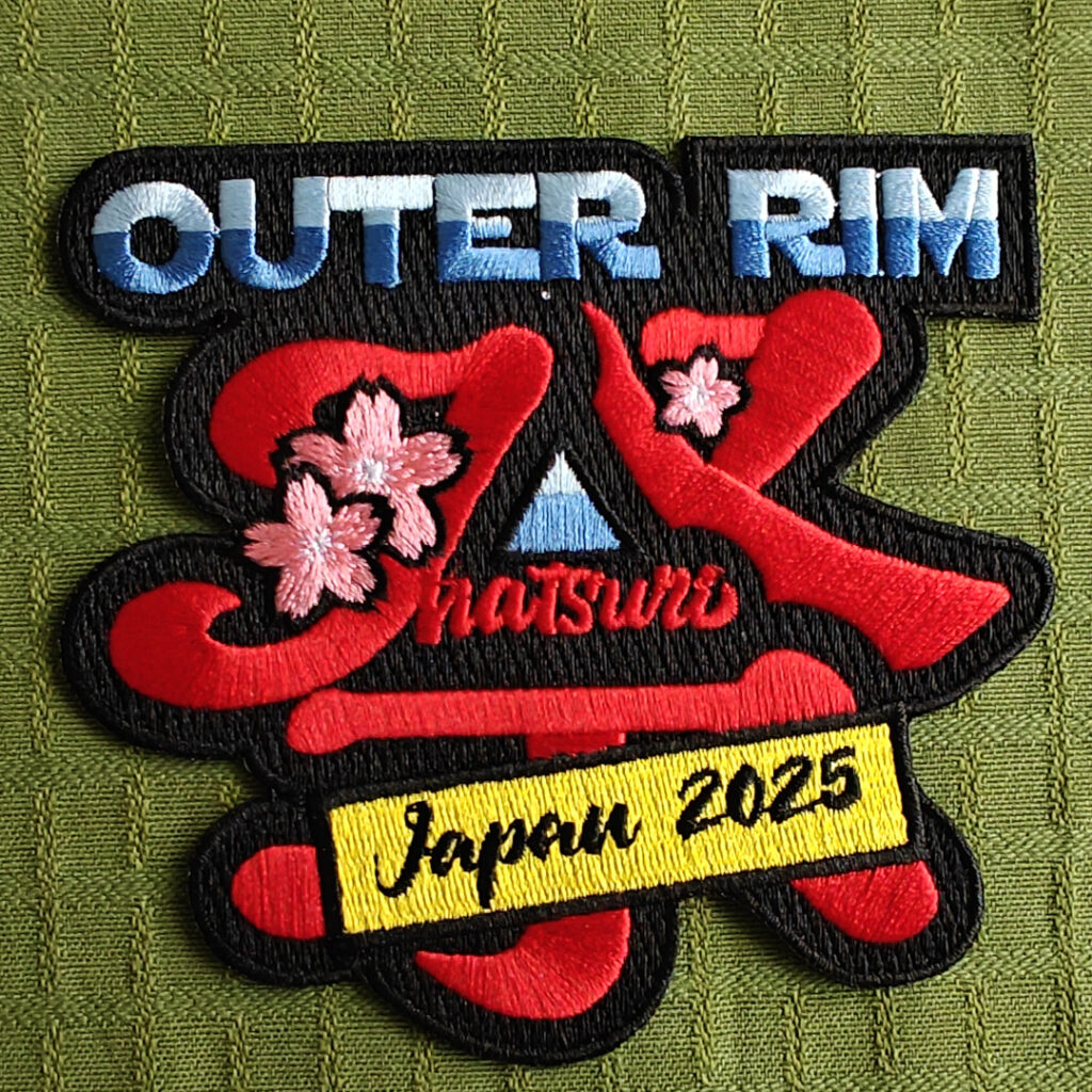 An embroidered patch with three lines of text.The top line is in blue: "Outer Rim". The middle line is a red, stylized japanese kanji with cherry blossoms, and also regular latin letters: "Matsuri".The bottom line is a yellow box with black letters: "Japan 2025".