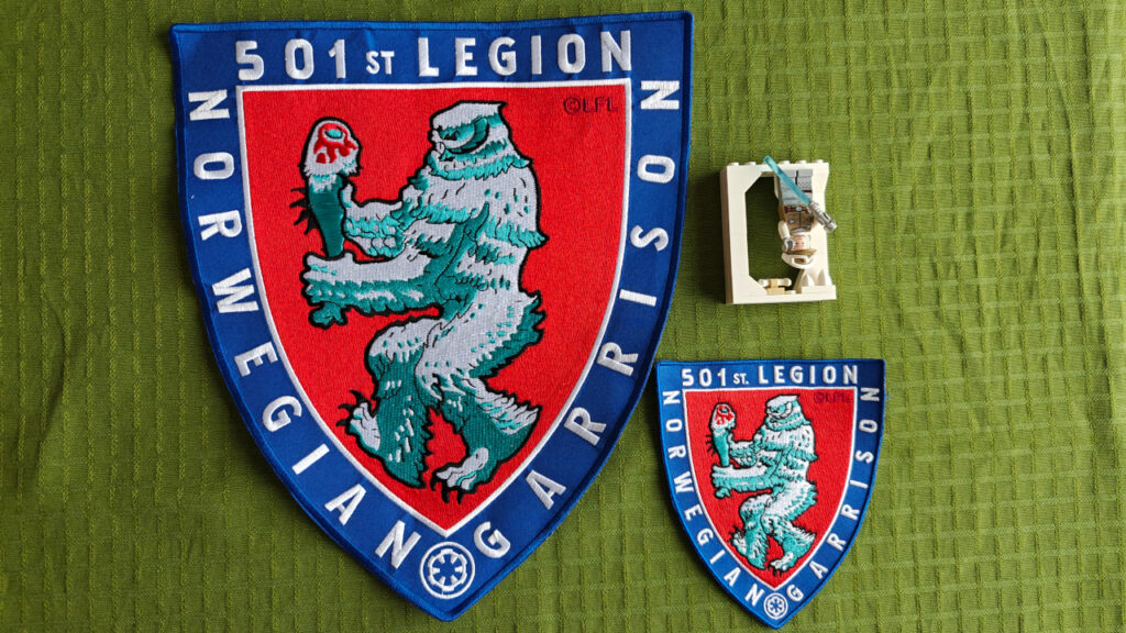 A embroidered patch in the shape of a shield, with a wampa carrying a taun-taun leg. The design is inspired by the Coat of Arms of Norway. A regular patch and a LEGO figure is also displayed for scale.