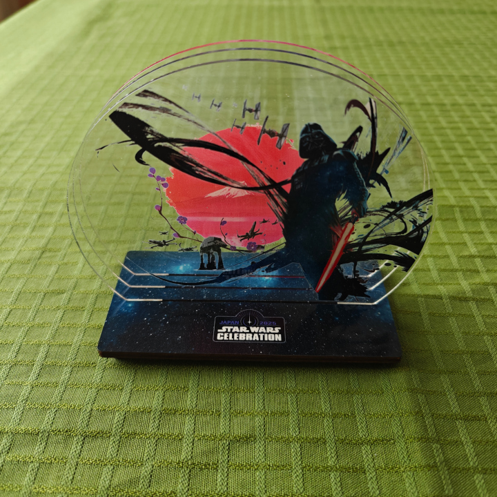 The Celebration key art as a sculpture. The base is black and blue and decorated with stars. It has three slots, each with a transparent acrylic disc with part of the key art, creating an image with depth.