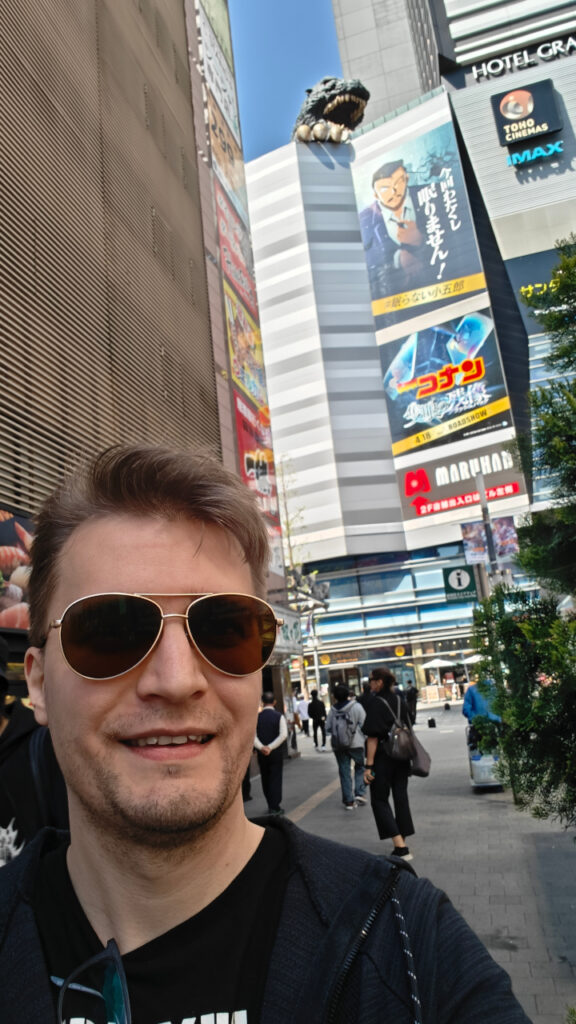 I am walking on the street. On one of the buildings behind me we see the head and claw of Gojira. Part of the building sign is seen: "Hotel Gra..."